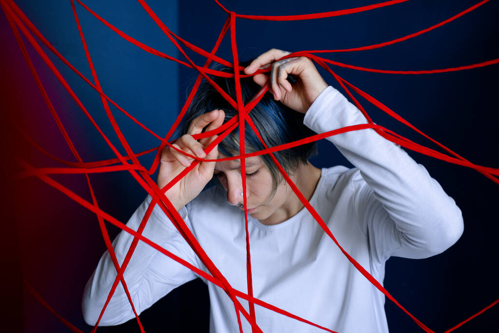 Young woman is entangled in red threads on blue walls background. Concept of obsessive states, paranoia, mental illness. - Photo, Image