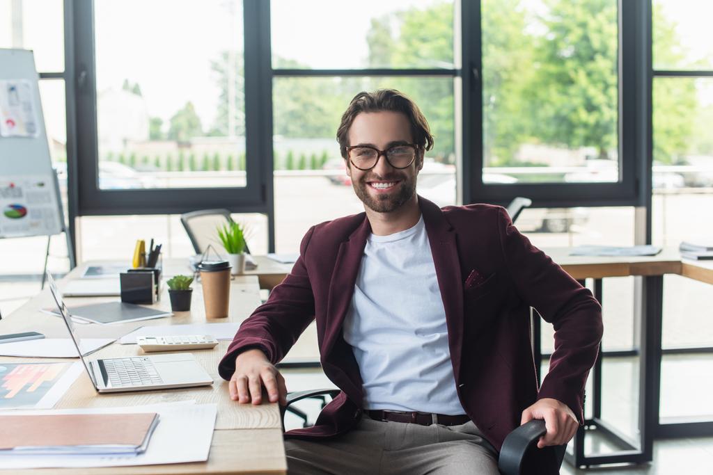 Positiver Geschäftsmann mit Brille, Kamera in der Nähe von Geräten, Taschenrechner und Papieren im Büro  - Foto, Bild
