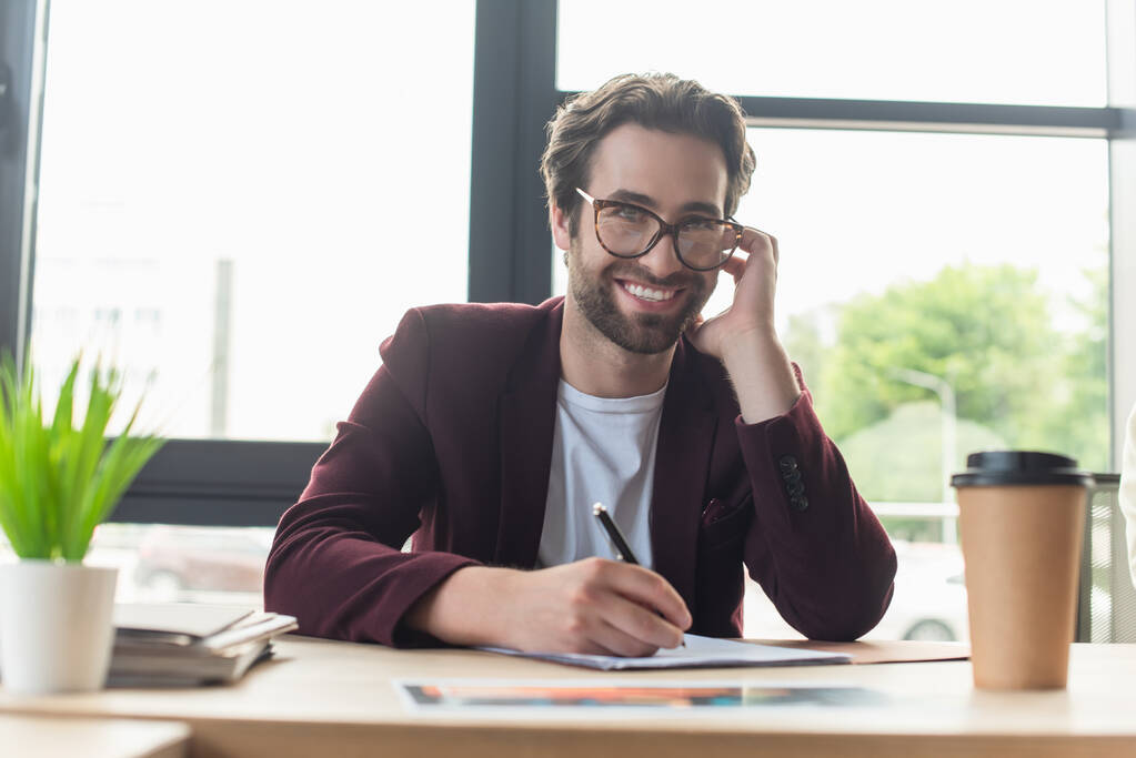 Позитивний бізнесмен в окулярах пише на папері біля розмитого виносного напою в офісі
  - Фото, зображення