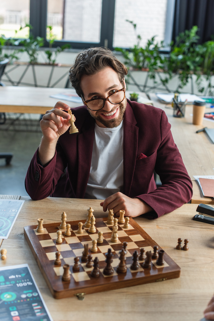 Uśmiechnięty biznesmen w okularach trzymający figurę szachową w biurze  - Zdjęcie, obraz