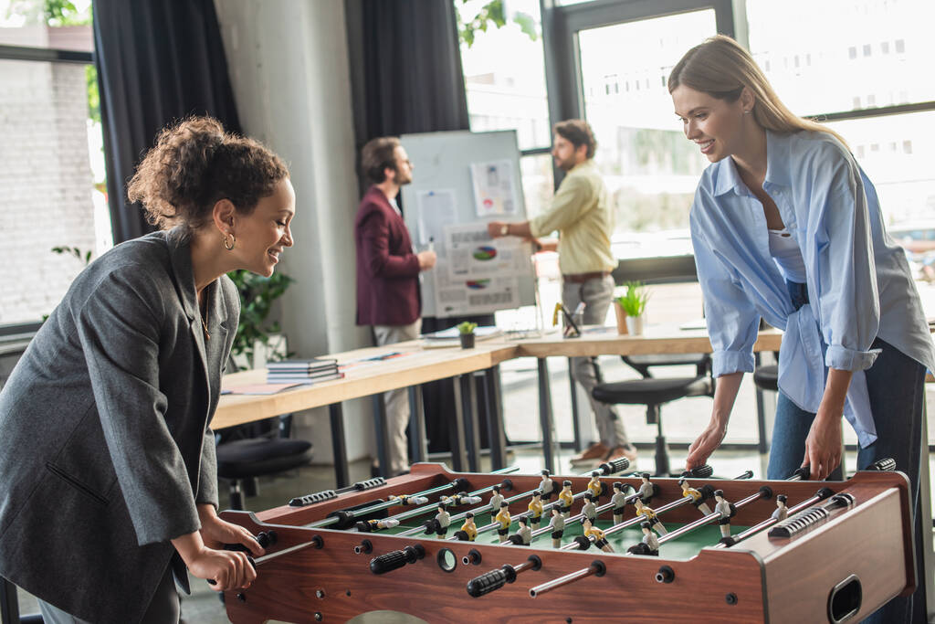 Mulheres de negócios inter-raciais alegres jogando futebol de mesa enquanto colegas desfocados trabalhando no escritório  - Foto, Imagem