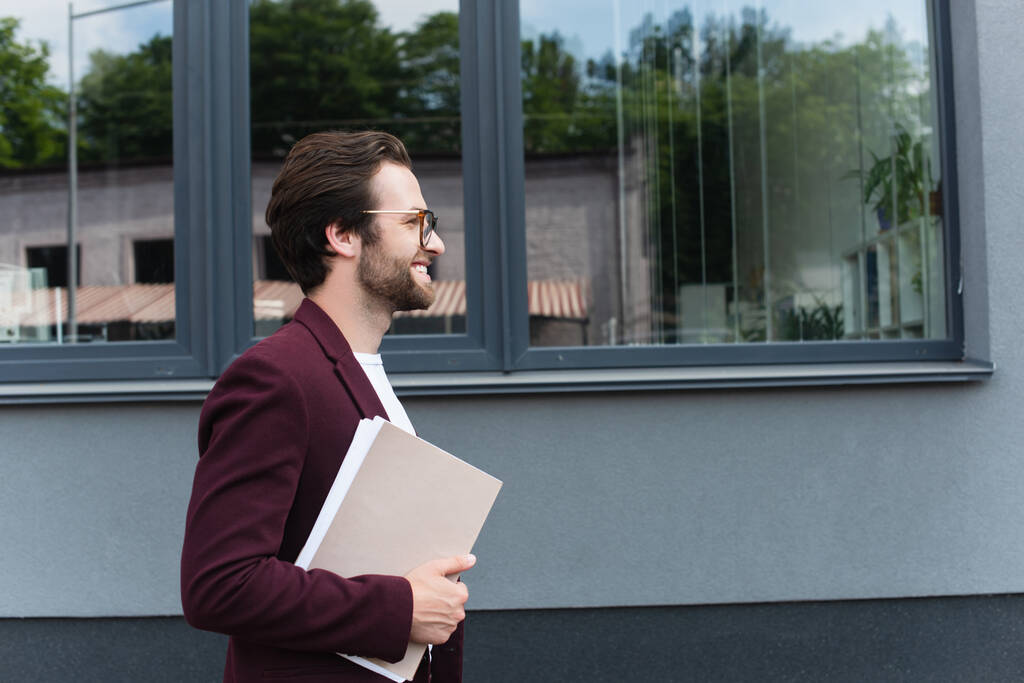 Πλευρική άποψη του χαμογελαστού επιχειρηματία με φακέλους χαρτί περπάτημα κοντά σε κτίριο σε εξωτερικούς χώρους  - Φωτογραφία, εικόνα