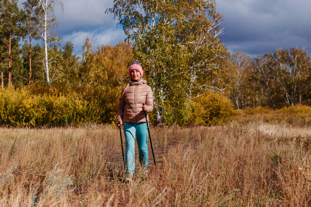 uma mulher idosa de sessenta anos de idade em sportswear está envolvida em caminhadas nórdicas com paus na floresta no outono - Foto, Imagem