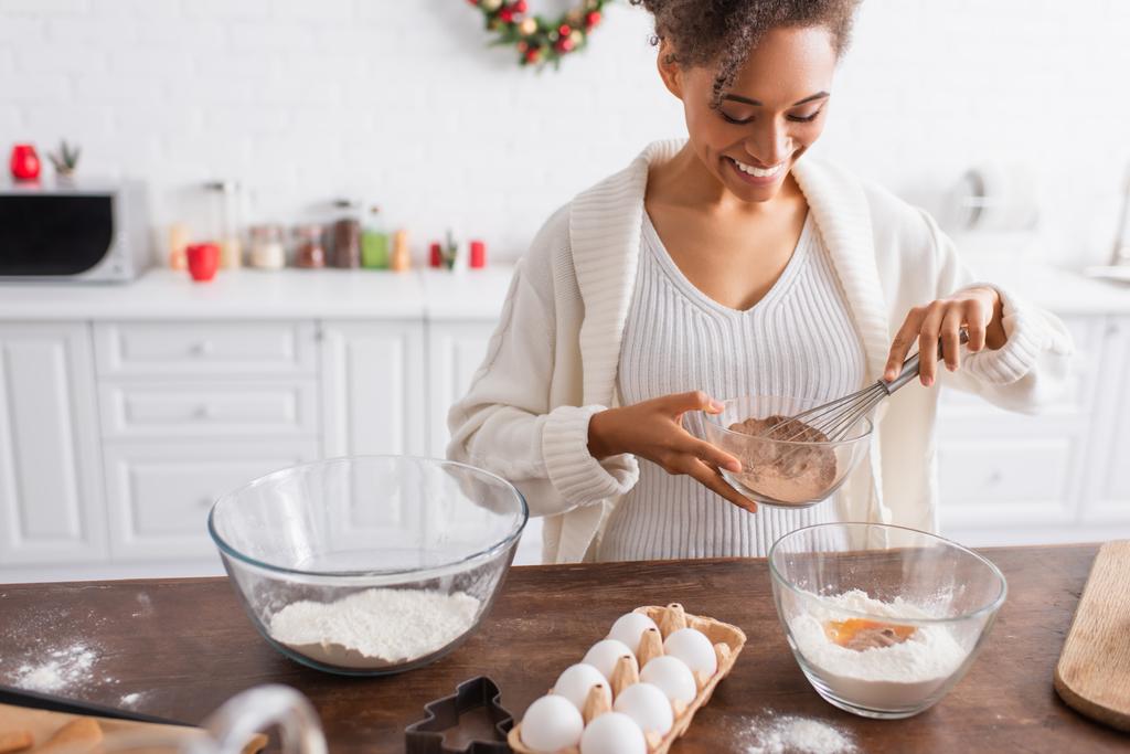 Mujer afroamericana sonriente sosteniendo cacao mientras cocina cerca de ingredientes y cortador de galletas en la cocina  - Foto, Imagen