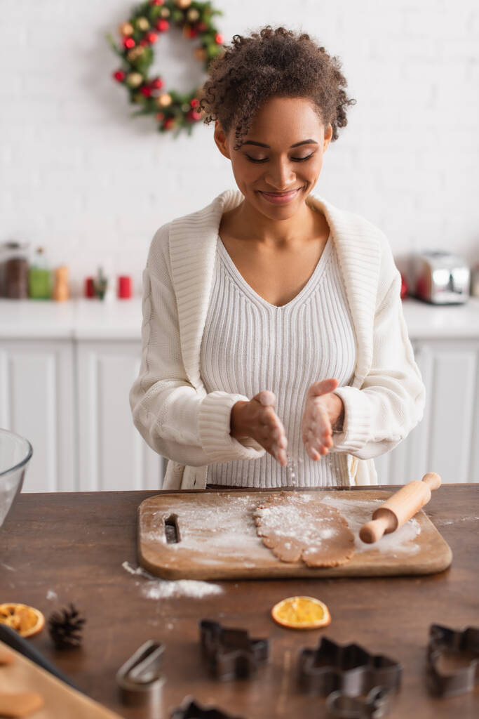 Усміхнена афроамериканка поливає борошно на тісто поблизу прокатного штифта та різаків для печива на кухні
  - Фото, зображення
