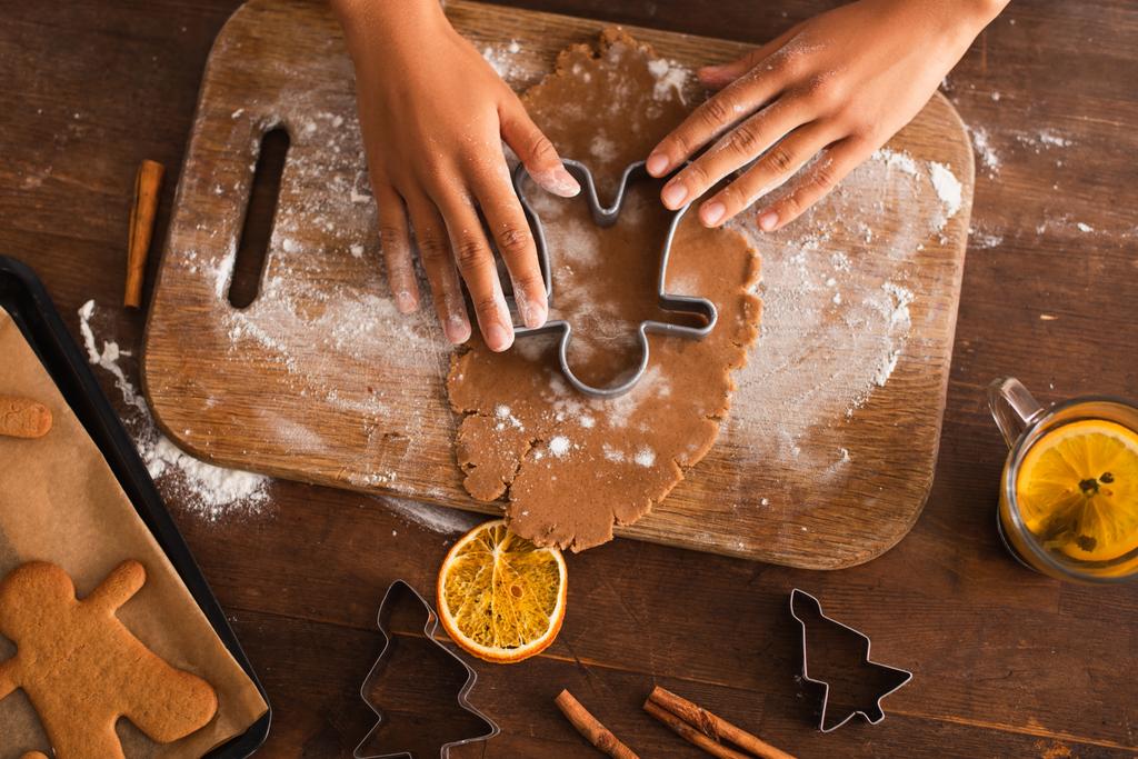 Vista superior de la mujer afroamericana presionando cortador de galletas de Navidad en la masa cerca de palitos de canela en la cocina  - Foto, imagen