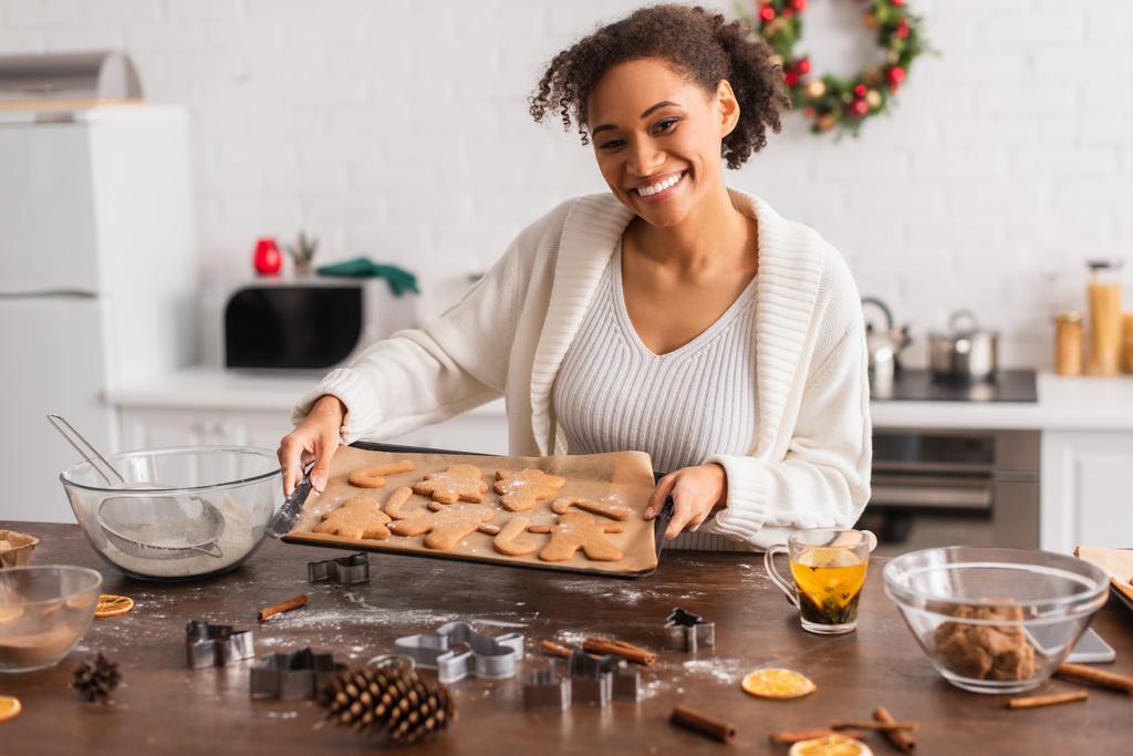 Mulher americana africana alegre segurando biscoitos de Natal na bandeja perto de ingredientes na cozinha  - Foto, Imagem