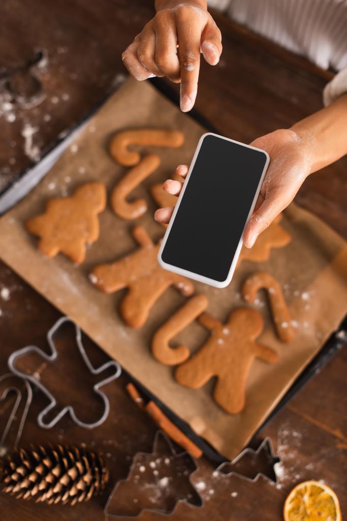 アフリカ系アメリカ人女性のトップビューの小麦粉とともに手にスマートフォンを保持近くぼやけたクリスマスクッキーでキッチン  - 写真・画像
