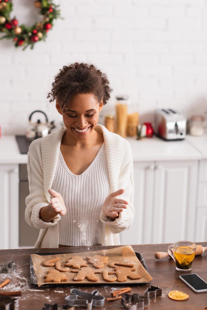 Sourire femme afro-américaine verser de la farine sur les biscuits de Noël près de smartphone et thé  - Photo, image