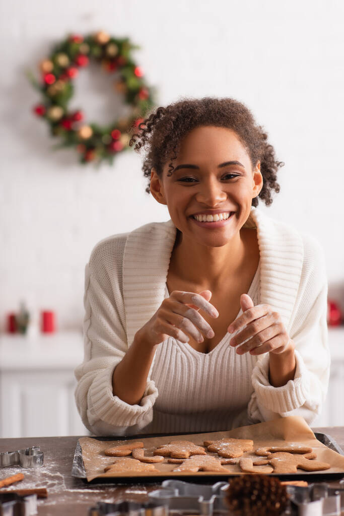 Positive afrikanisch-amerikanische Frau blickt in die Kamera fast Weihnachtsplätzchen in der Küche  - Foto, Bild