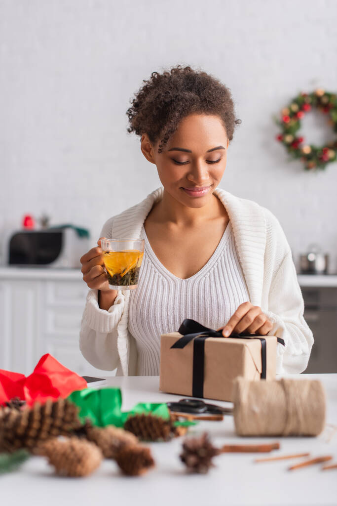 Young african american woman holding tea while decorating christmas gift at home  - Photo, Image