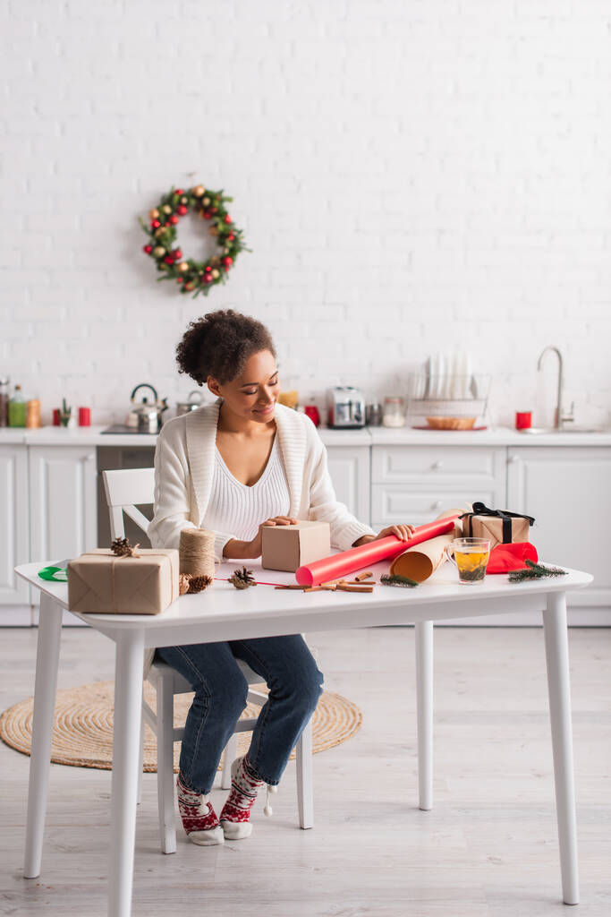 Sorridente donna afro-americana in cerca di scatola regalo e decorazioni natalizie a casa  - Foto, immagini