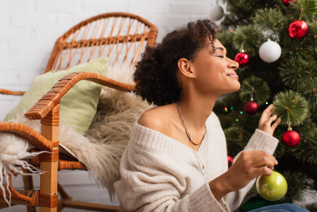 Vista lateral da mulher americana africana alegre segurando bola de Natal perto de árvore e cadeira em casa  - Foto, Imagem