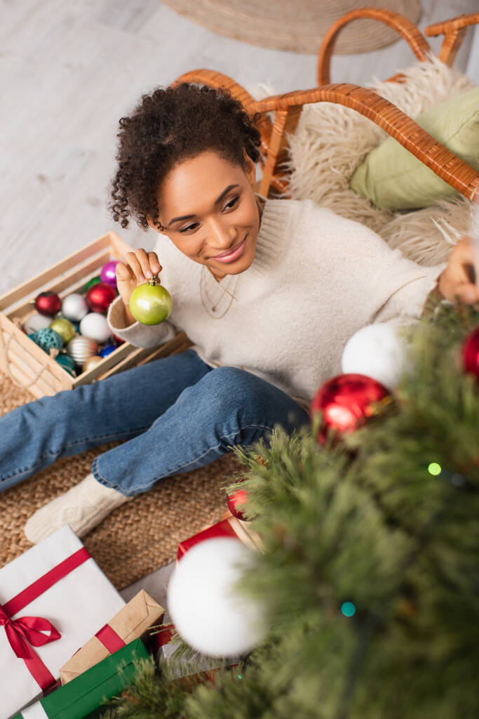 Vista de ángulo alto de la sonriente mujer afroamericana sosteniendo la pelota cerca del borroso árbol de navidad y regalos en casa  - Foto, imagen