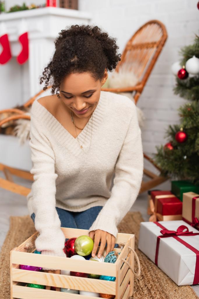 Afroamerykanka patrzy na pudełko z kulkami świątecznymi w pobliżu prezentów w domu  - Zdjęcie, obraz