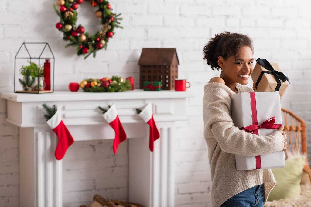 Veselý africký Američan žena drží vánoční dárky v blízkosti rozmazané krb doma  - Fotografie, Obrázek