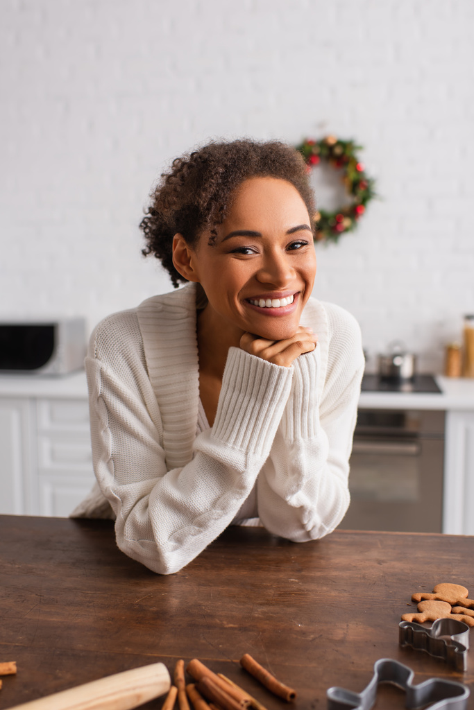 Χαρούμενη Αφροαμερικανίδα που κοιτάζει κάμερα κοντά σε ξυλάκια κανέλας και κόφτες μπισκότων στην κουζίνα.  - Φωτογραφία, εικόνα