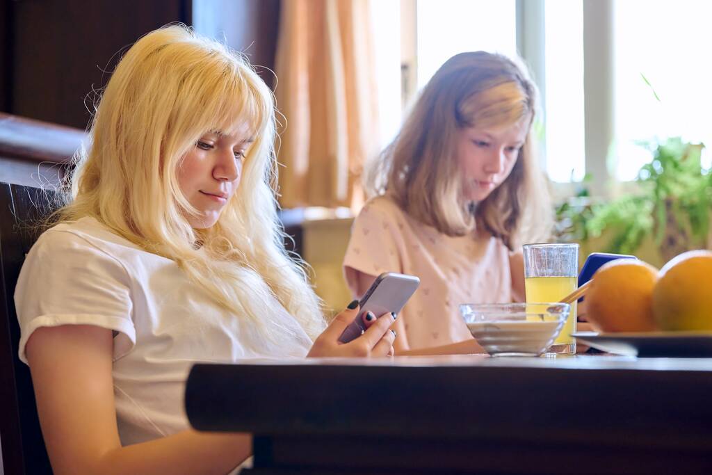 Παιδιά δύο αδελφές κορίτσια τρώνε στο σπίτι, κοιτάζοντας smartphones - Φωτογραφία, εικόνα