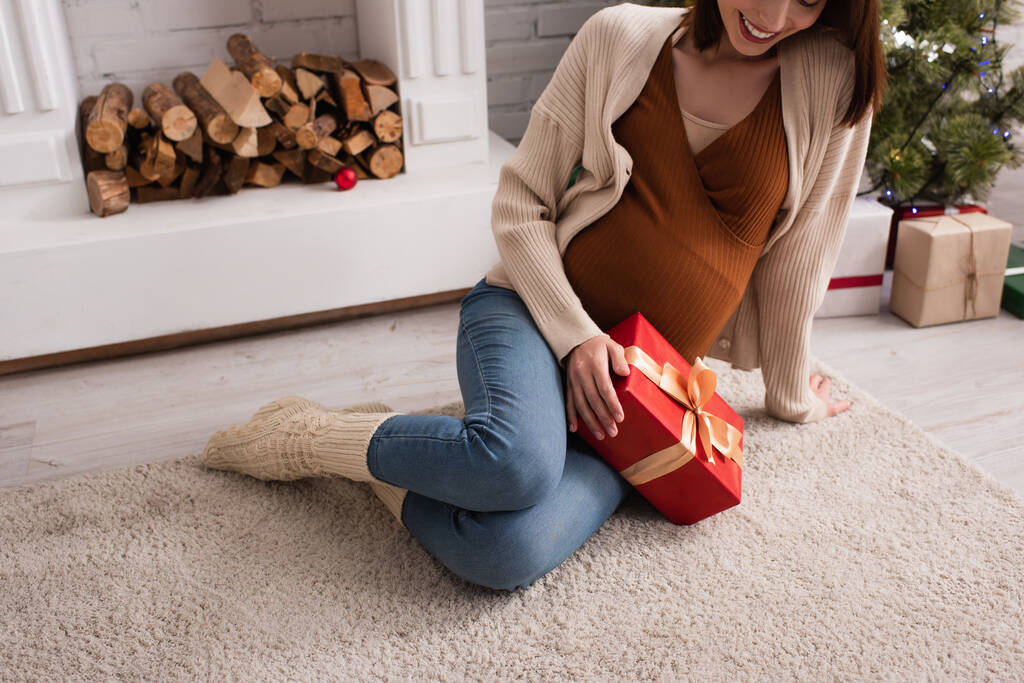 Gülümseyen hamile bir kadının şöminenin yanında noel hediyesiyle oturması. - Fotoğraf, Görsel