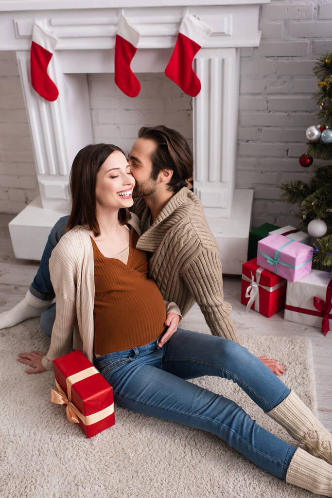 ギフトボックスの近くの床に座って暖炉を飾る夫と幸せな妊婦 - 写真・画像