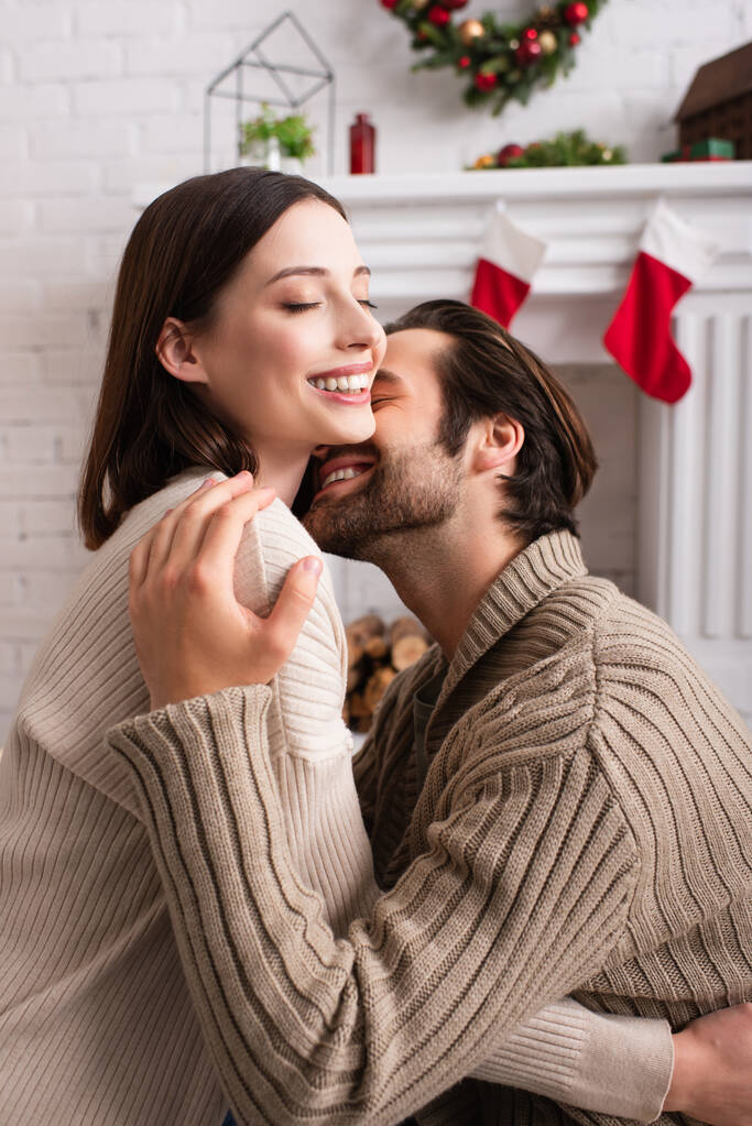 задоволений чоловік обіймає дружину біля розмитого каміна з різдвяною прикрасою
 - Фото, зображення
