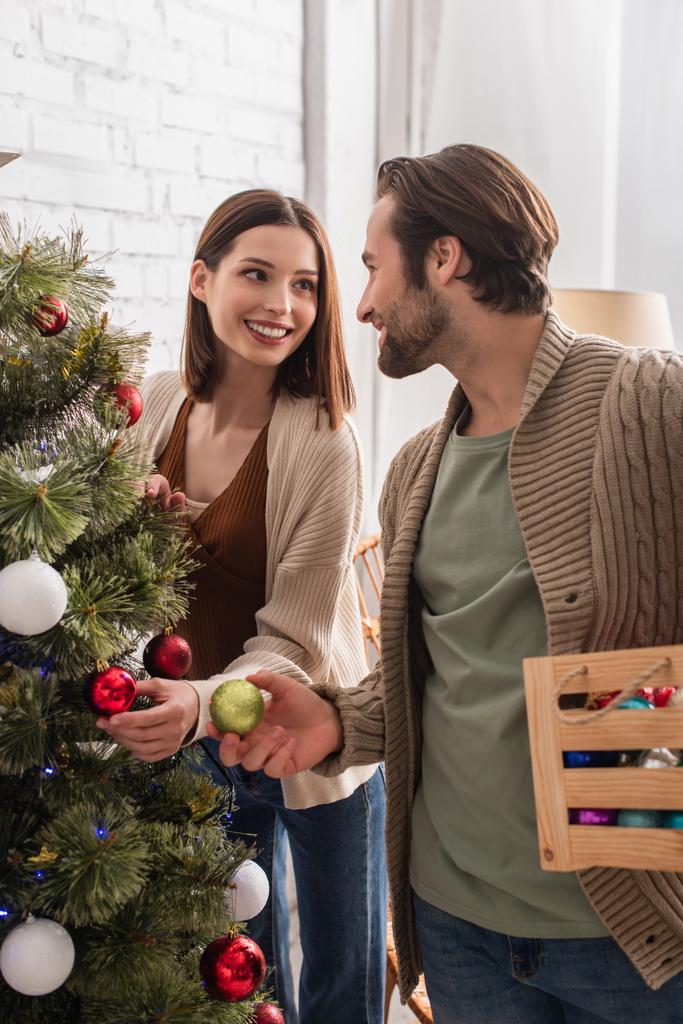 freudiges Paar lächelt sich beim Schmücken des Weihnachtsbaums zu Hause an - Foto, Bild