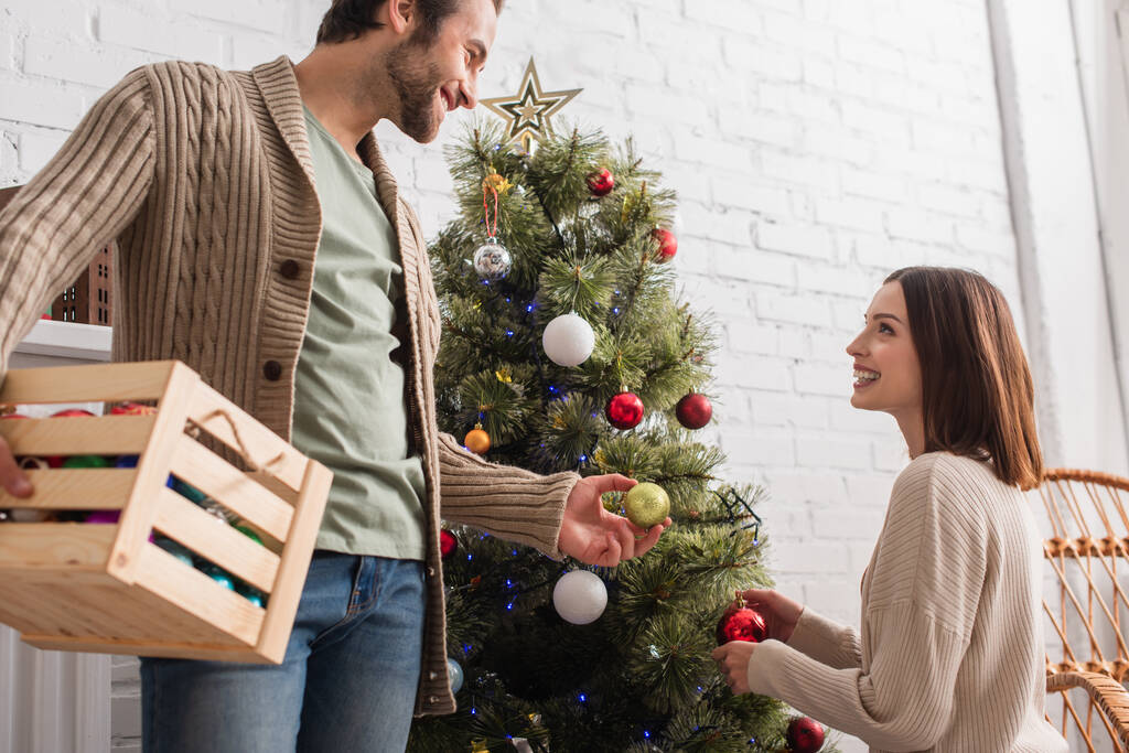 uomo sorridente con scatola di legno dando palla di Natale alla moglie decorare pino in soggiorno - Foto, immagini