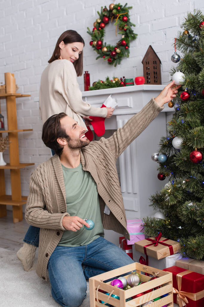 Gülümseyen bir adam çam ağacını süslüyor ve bulanık bir eş elinde şöminenin yanındaki Noel çorabını tutuyor. - Fotoğraf, Görsel