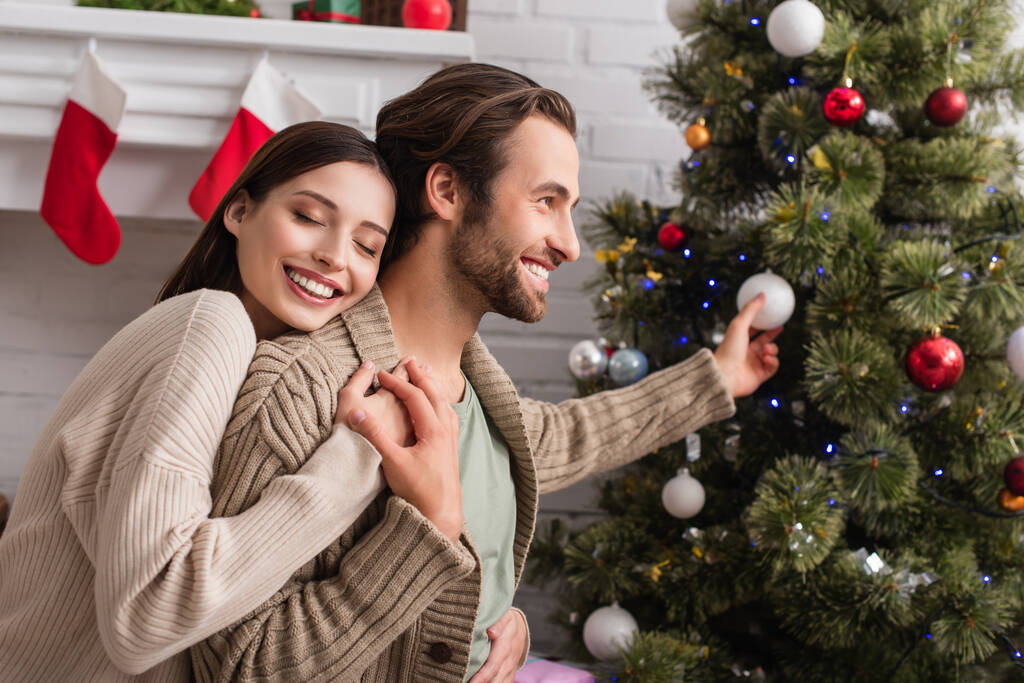 Ευτυχισμένη γυναίκα με κλειστά μάτια αγκαλιάζει χαμογελαστό σύζυγο διακοσμώντας θολό χριστουγεννιάτικο δέντρο - Φωτογραφία, εικόνα