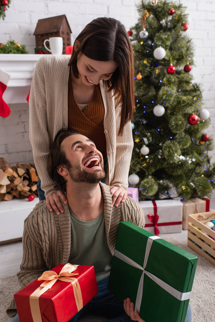 здивований чоловік тримає подарунки біля дружини і розмиту ялинку у вітальні
 - Фото, зображення