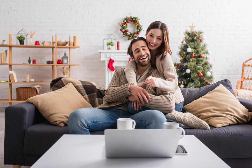 Mutlu bir kadın kocasını kucaklıyor ve oturma odasındaki kanepede dizüstü bilgisayarını işaret ediyor. - Fotoğraf, Görsel