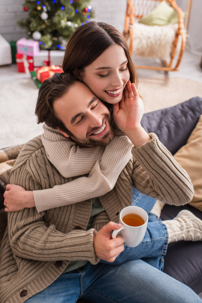 hombre complacido con taza de té tocando la cara de la esposa abrazándolo en la sala de estar con decoración de Navidad - Foto, Imagen