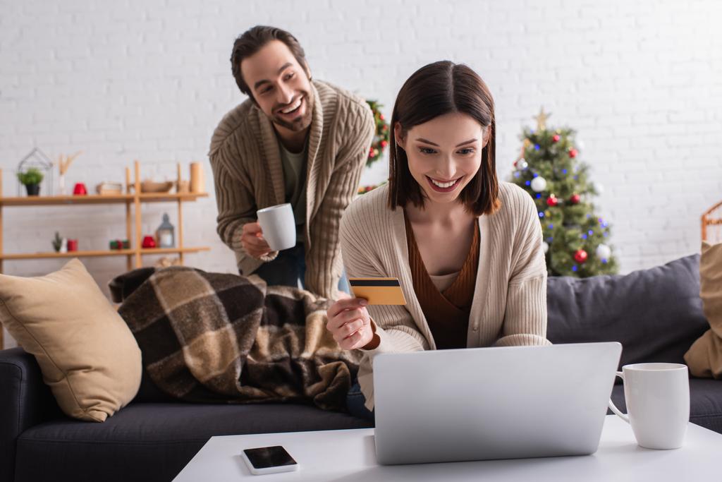 sonriente mujer con tarjeta de crédito utilizando el ordenador portátil cerca borrosa marido con taza de té - Foto, Imagen
