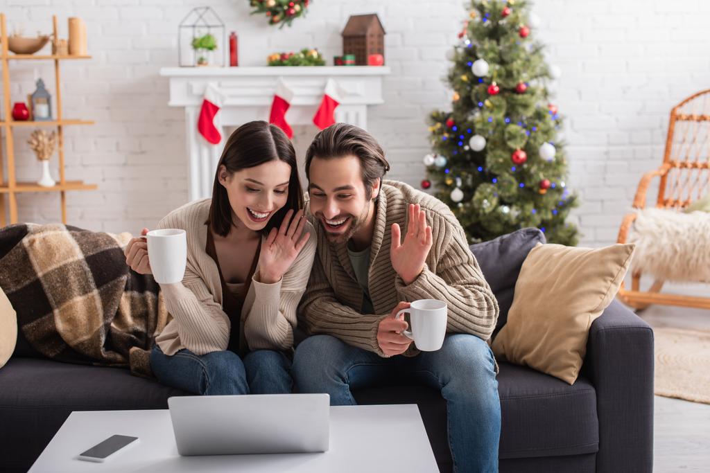 fröhliches Paar mit Teetassen, winkenden Händen bei Videoanruf im dekorierten Wohnzimmer - Foto, Bild