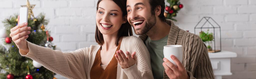 felice donna prendendo selfie con marito in possesso di una tazza di tè in soggiorno con decorazione natalizia, banner - Foto, immagini