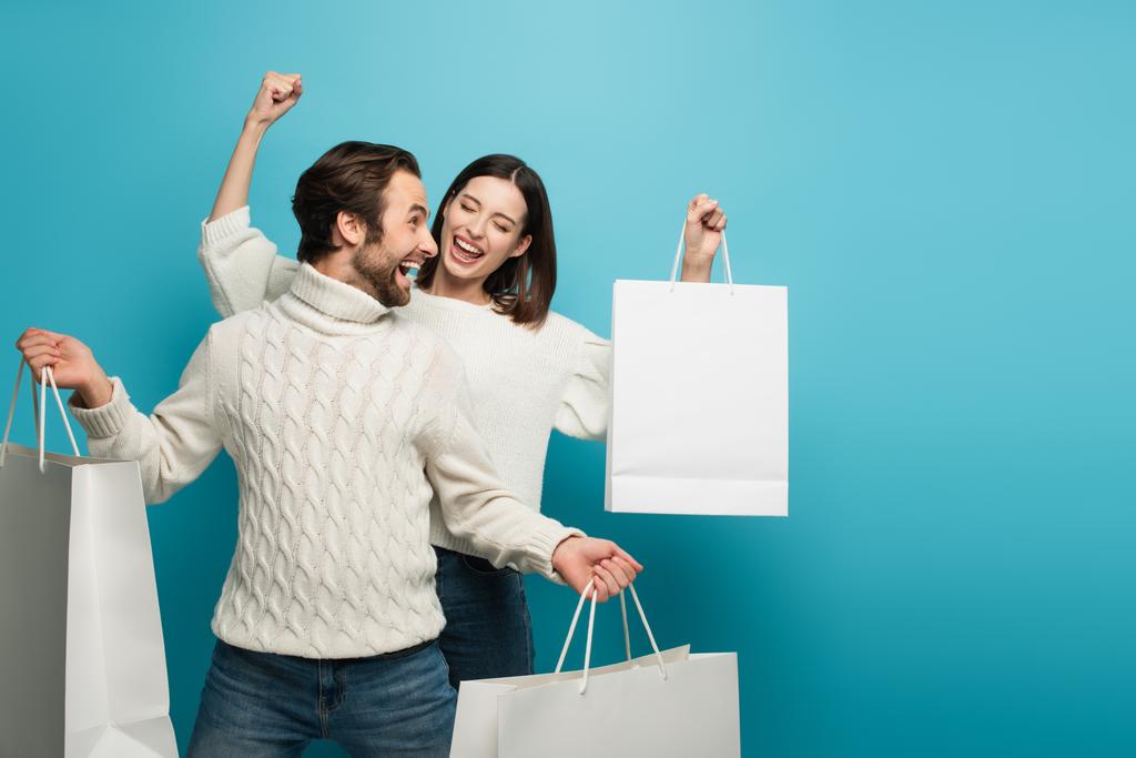 fröhliche Frau hält Einkaufstasche in der Hand und zeigt Erfolgsgeste neben Mann mit Einkäufen auf blau - Foto, Bild
