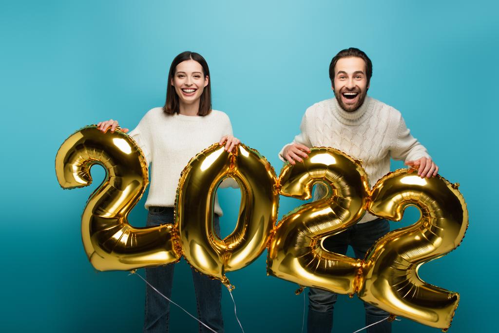 cheerful couple looking at camera while holding golden balloons in shape of 2022 on blue - Photo, Image