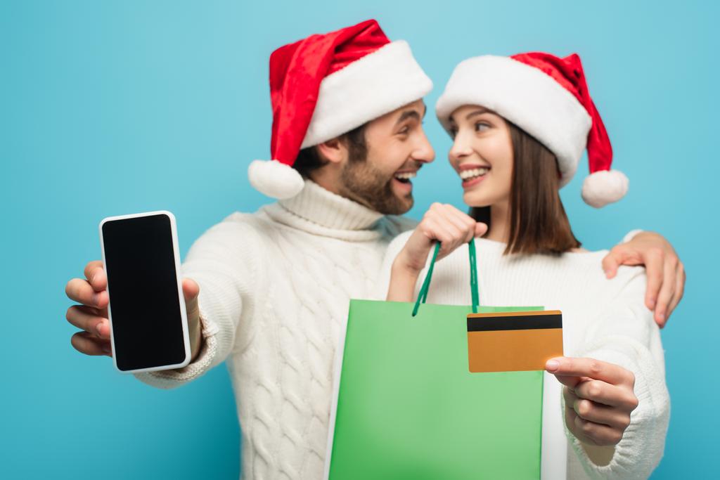 pareja borrosa en sombreros de santa sonriendo el uno al otro mientras sostiene el teléfono inteligente, tarjeta de crédito y bolsas de compras aisladas en azul - Foto, Imagen