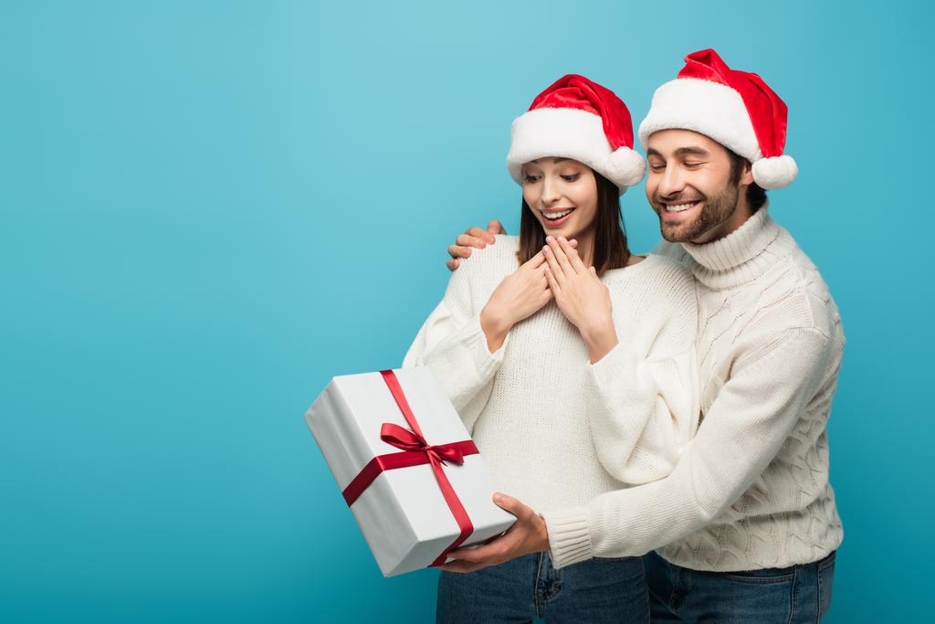 χαμογελαστός άντρας με καπέλο Σάντα κρατώντας χριστουγεννιάτικο δώρο κοντά κατάπληκτη γυναίκα απομονωμένη στο μπλε - Φωτογραφία, εικόνα