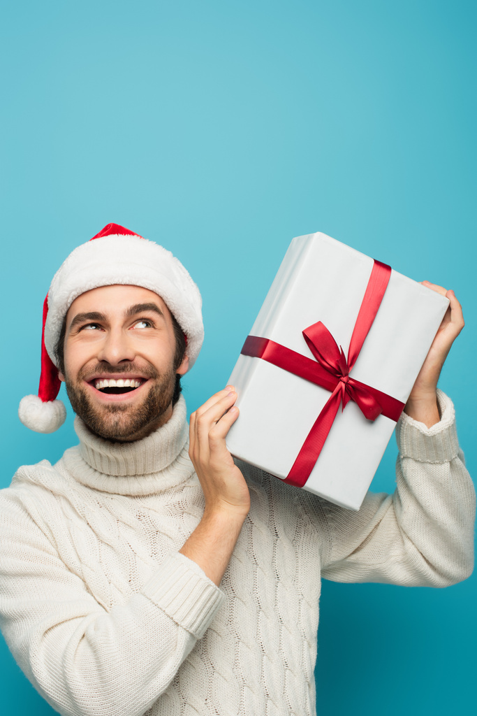 homme heureux et curieux dans santa chapeau secouant boîte cadeau isolé sur bleu - Photo, image