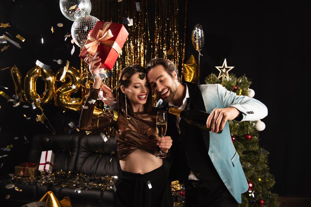 homem feliz derramando champanhe enquanto celebra o Natal com mulher elegante no preto - Foto, Imagem