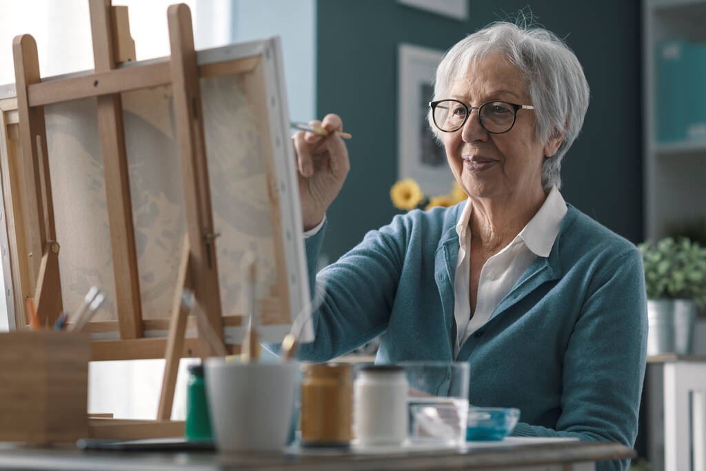 Gelukkig senior vrouw zitten aan het bureau thuis en schilderen op doek - Foto, afbeelding