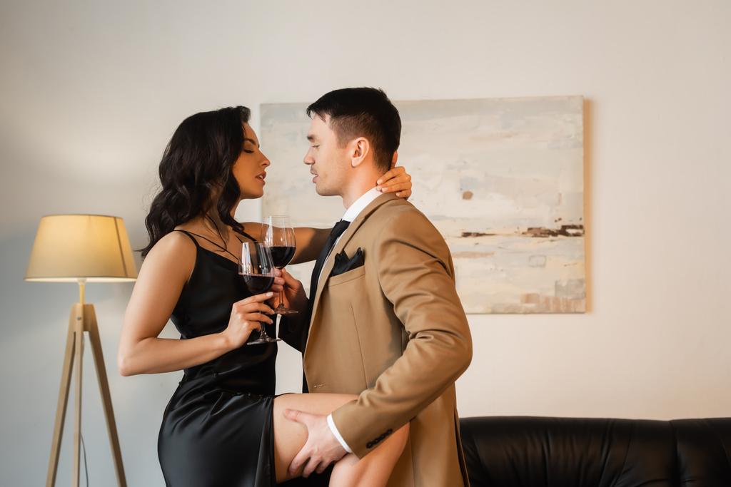 vista lateral del hombre levantando pierna de mujer seductora en vestido de deslizamiento con copa de vino tinto  - Foto, Imagen