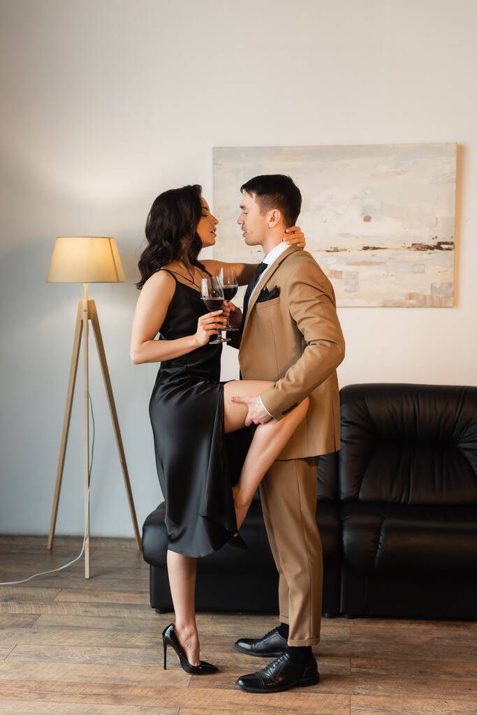 comprimento total do homem apaixonado levantando perna de mulher sedutora em vestido de deslizamento com copo de vinho tinto  - Foto, Imagem