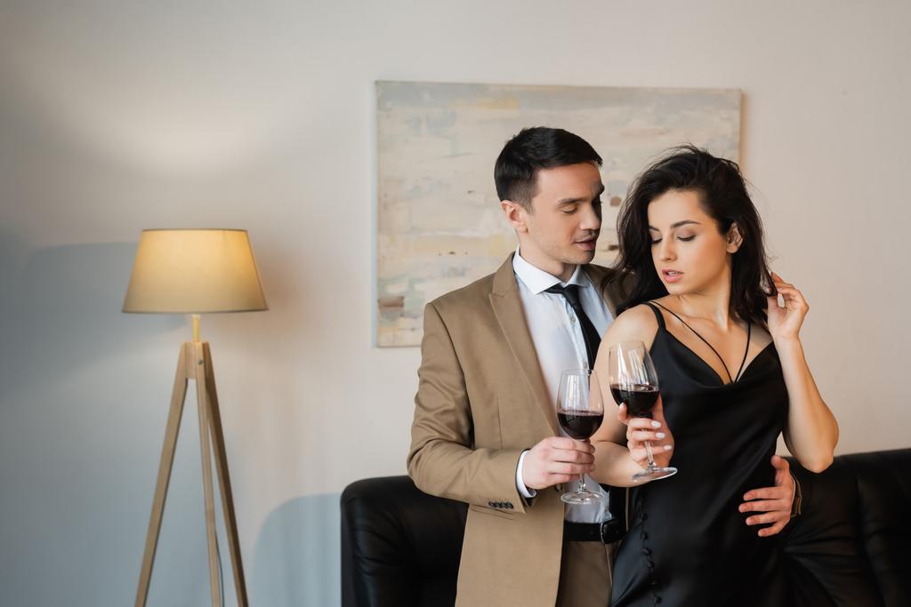 若いセクシーなカップルがリビングルームで赤ワインのグラスを持ち - 写真・画像