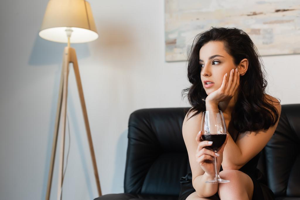 mujer bonita en vestido negro deslizante sosteniendo copa de vino y sentado en el sofá - Foto, imagen
