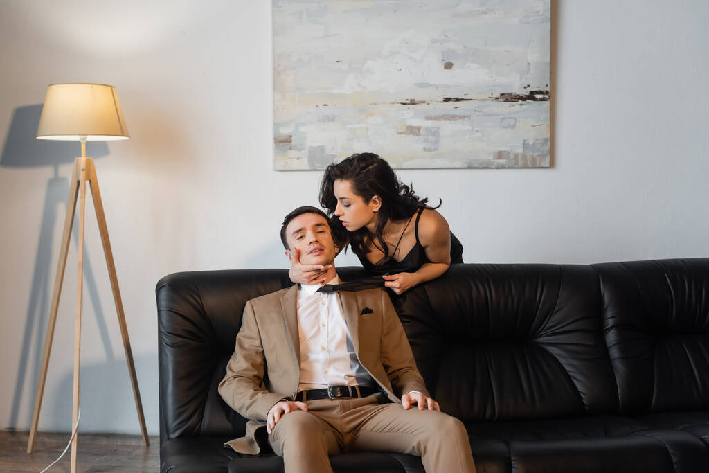 brunette nainen musta lipsahdus mekko vetämällä solmio kun viettelee mies sohvalla  - Valokuva, kuva