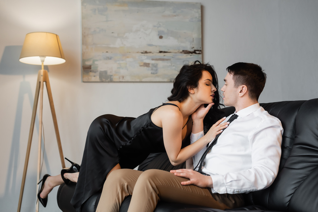 vista lateral de sexy jovem mulher em vestido de deslizamento preto seduzindo namorado no sofá  - Foto, Imagem