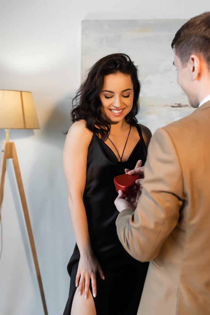 homályos férfi kezében szív alakú ajándék doboz közelében mosolygós barátnő fekete slip ruha  - Fotó, kép
