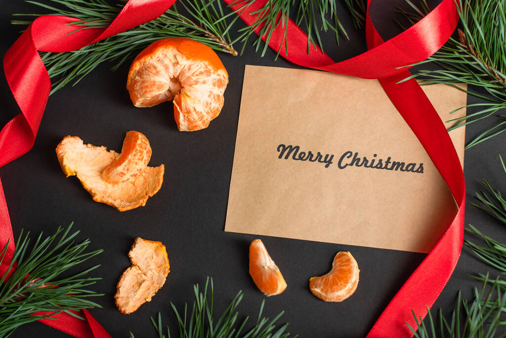 vue du dessus du papier artisanal avec joyeux lettrage de Noël près de mandarines pelées et branches de sapin sur noir - Photo, image